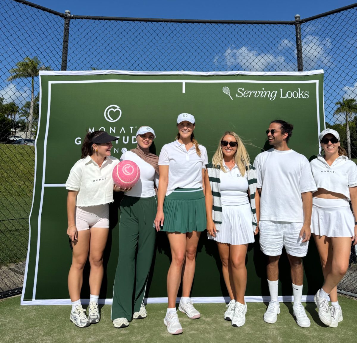 6 smiling people standing in front of a green tennis themed backdrop that says "Serving looks". One person is holding a giant pink sparkly tennis ball.